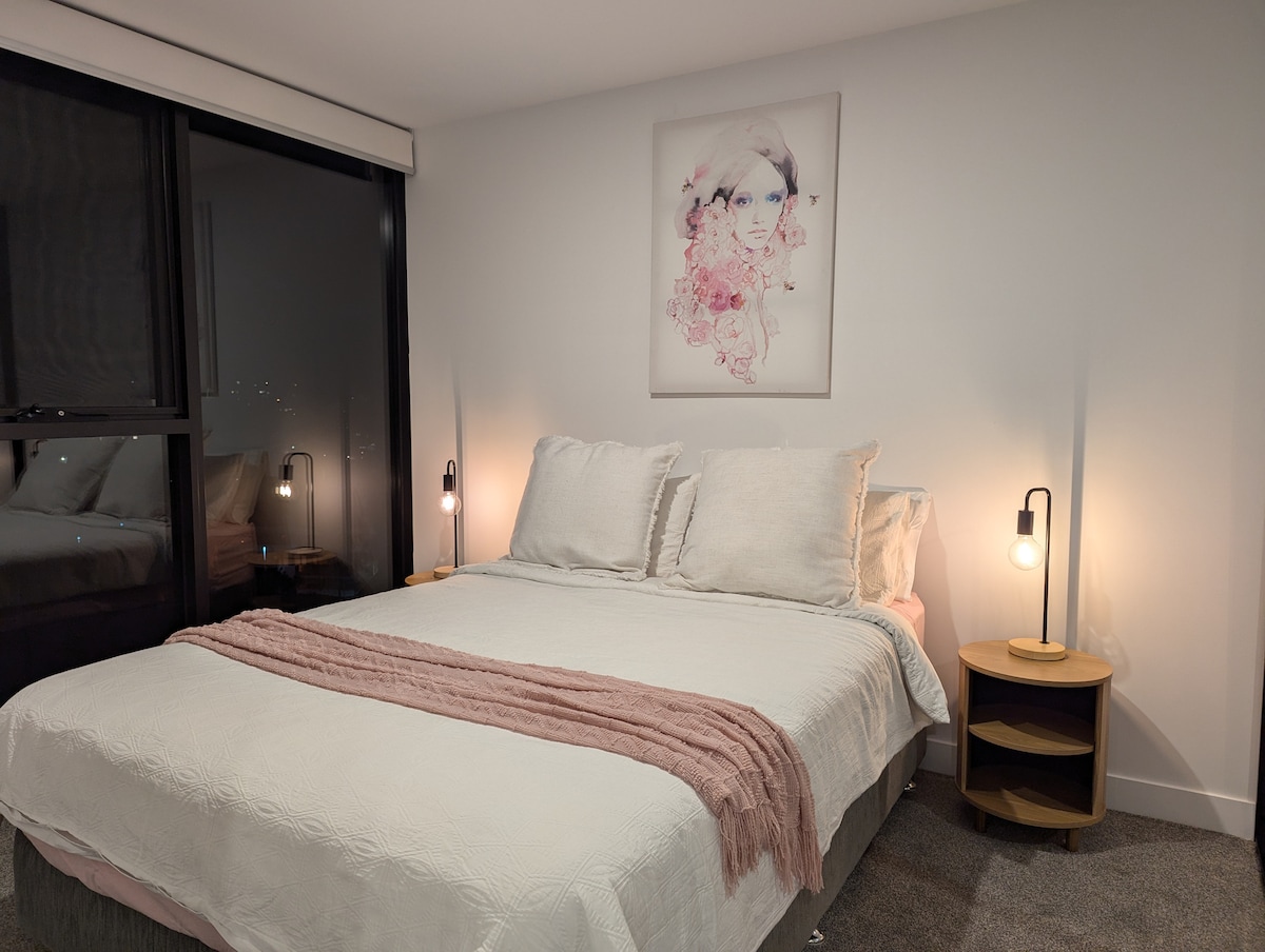 A well-appointed bedroom features a spacious bed dressed in white linens and soft pillows. Elegant bedside lamps are positioned on wooden tables, and a piece of wall art enhances the ambiance. Large windows provide natural light and a view of the surroundings.