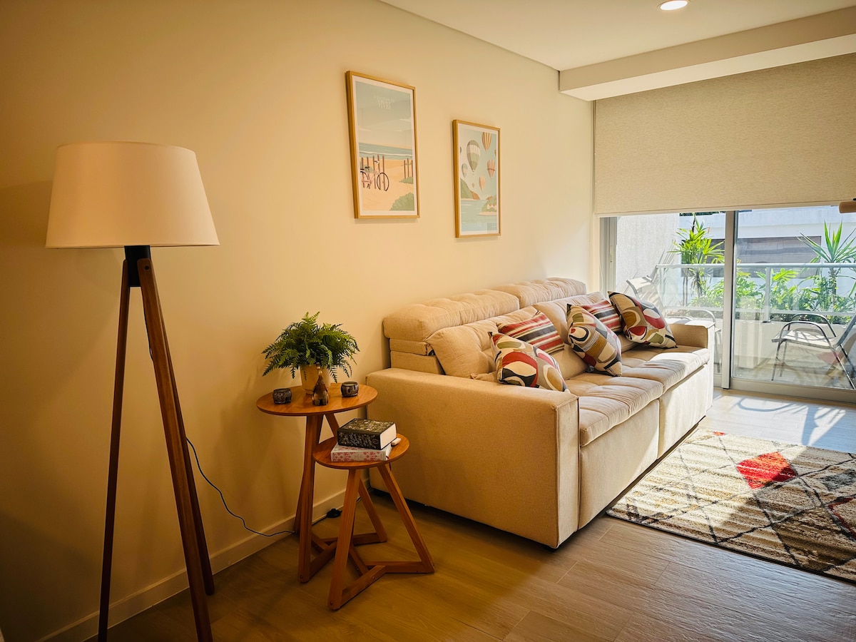 A light-filled living area features a comfortable sofa adorned with decorative pillows. A stylish floor lamp stands beside a small side table holding a plant and a book. Artwork is displayed on the wall above the sofa, enhancing the cozy atmosphere.