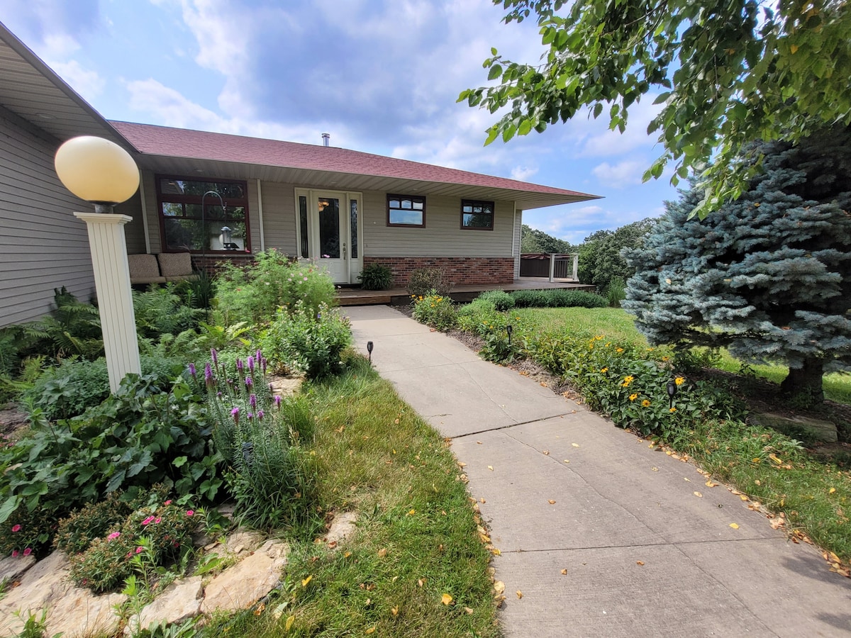 A well-maintained pathway leads to a welcoming entrance of the home, bordered by lush greenery and colorful flowers. The front porch, featuring comfortable seating, is shaded by a large overhang, while a lamp post adds gentle illumination to the landscaped yard.