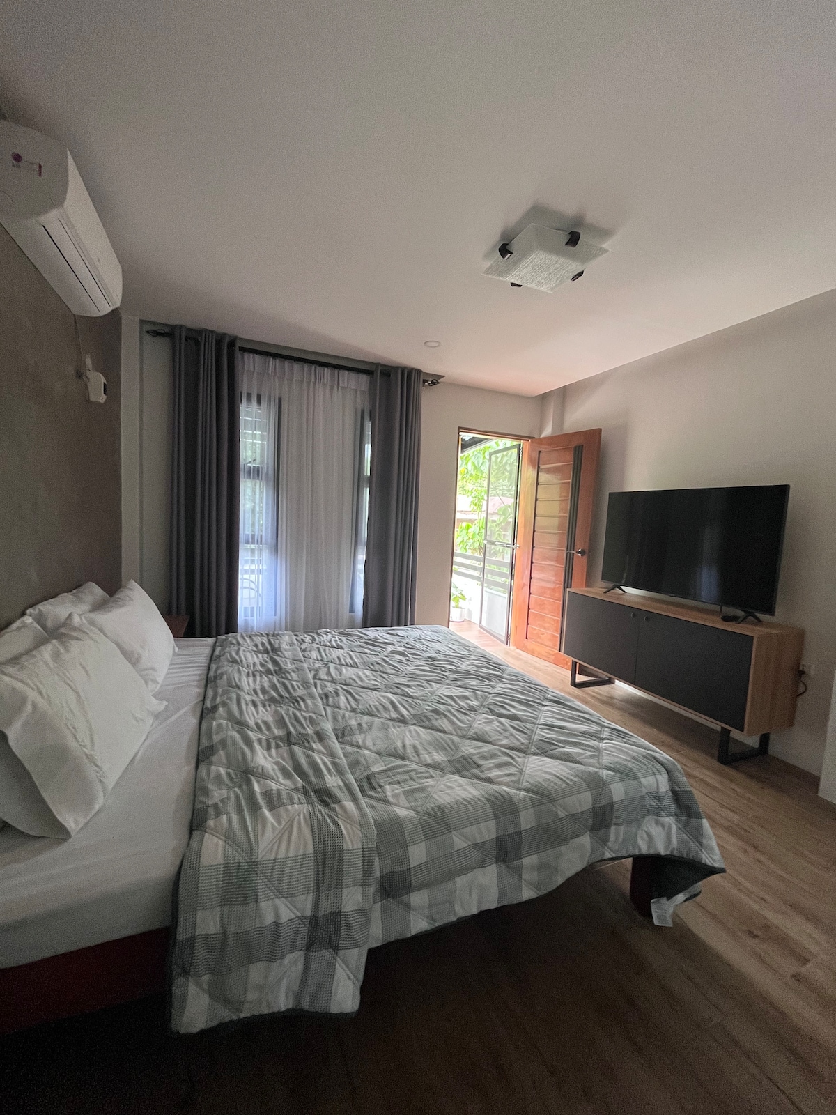 A spacious bedroom features a comfortable bed with a light grey quilt. Natural light filters through sheer curtains beside the window, which opens to an outdoor view. A flat-screen TV is positioned on a modern stand, and an air conditioner is mounted above the bed.