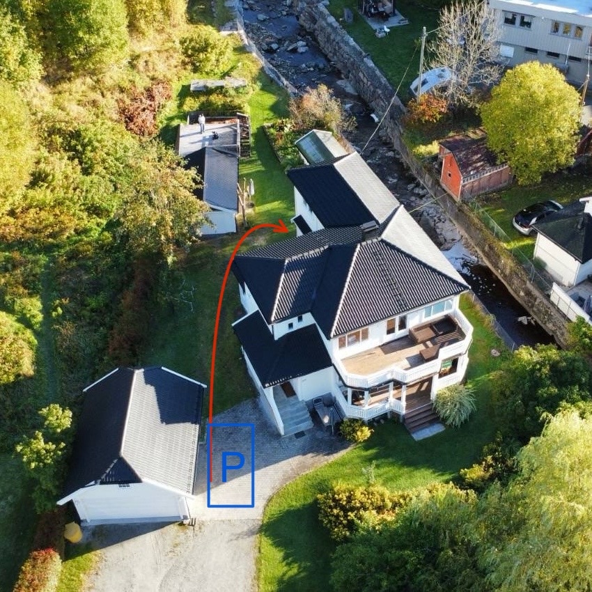 An aerial view of the property is presented, highlighting the main house and the adjacent structure containing the private apartment. A clearly marked area indicates guest parking, located near the entrance. Lush greenery and a flowing river are visible, contributing to the serene surroundings.
