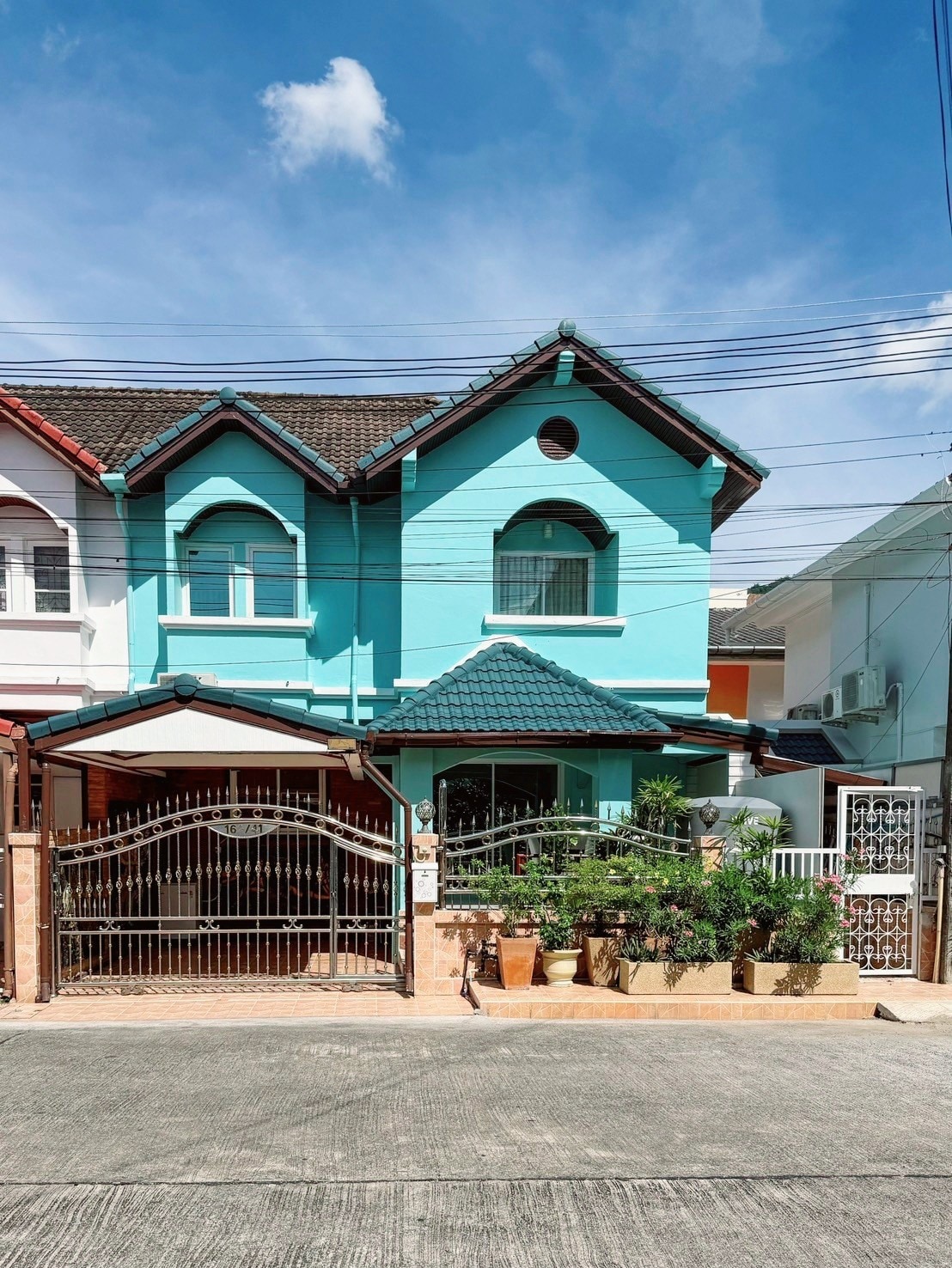 A charming two-story house is displayed, characterized by its vibrant turquoise exterior. The entrance is framed by a decorative fence, with potted plants adorning the pathway. Bright blue skies and wispy clouds create a serene backdrop, contributing to the welcoming atmosphere.
