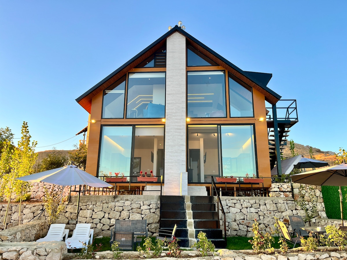 A modern two-story building features large glass windows, allowing natural light to illuminate the interior. The exterior incorporates wood and stone elements. A landscaped area with lounge chairs and umbrellas is visible in the foreground, complementing the natural surroundings.