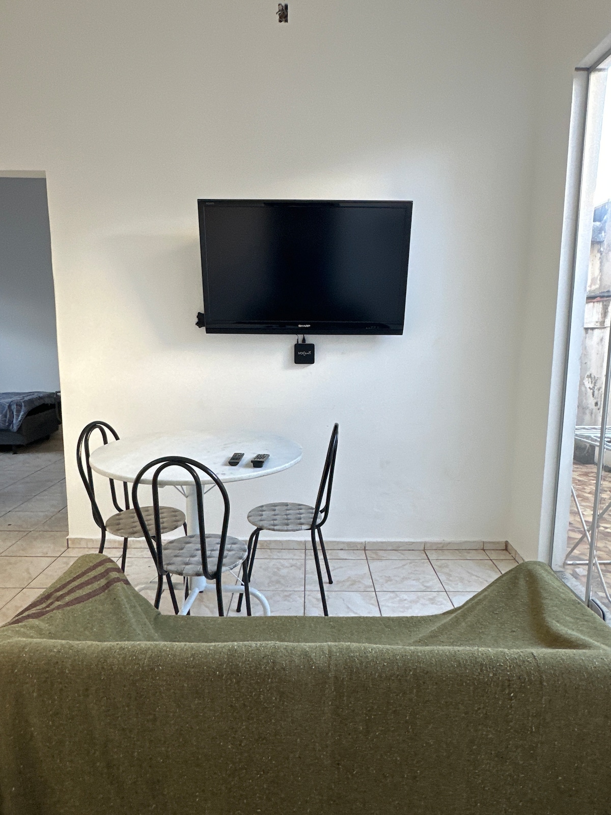A small dining area features a round table with three metal chairs. A flat-screen television is mounted on the wall opposite a sofa, partially covered with a green blanket. Natural light is entering through a nearby window.