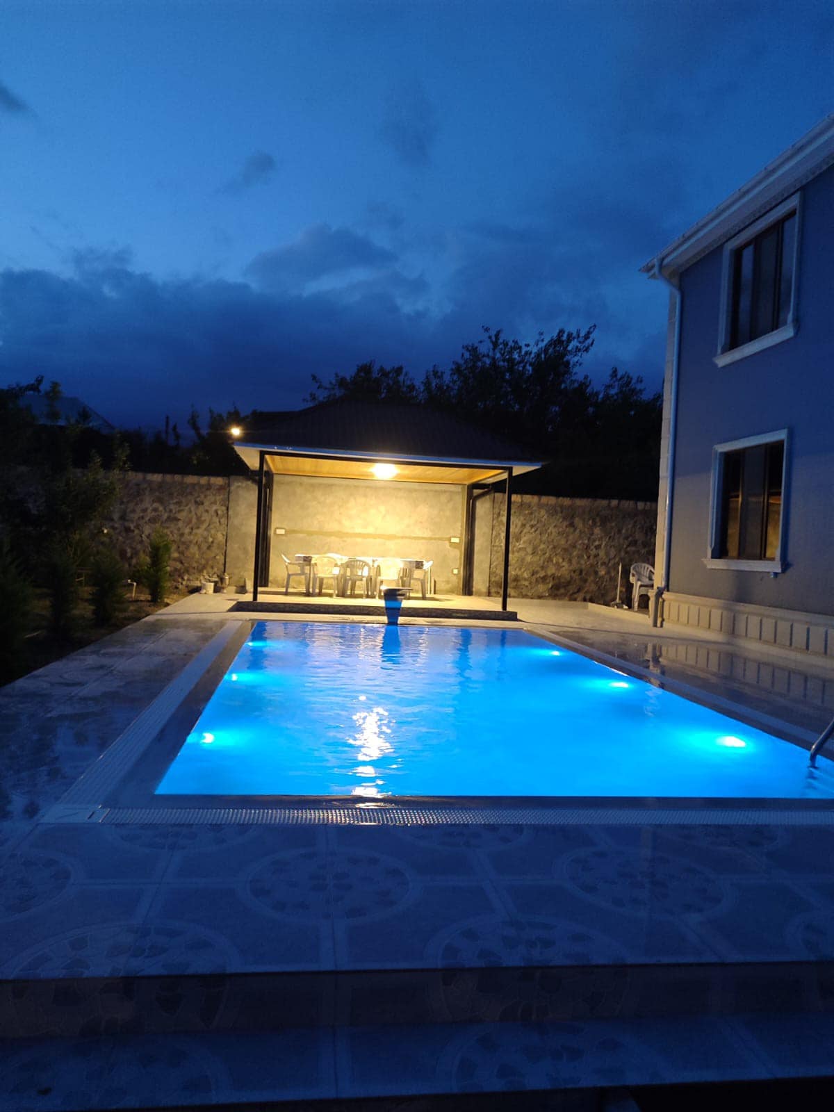 A serene pool area is illuminated by bluish lights under a clear night sky. The pool's shimmering surface reflects the surrounding greenery. A shaded seating area with several chairs is positioned nearby, providing a space for relaxation and enjoyment.