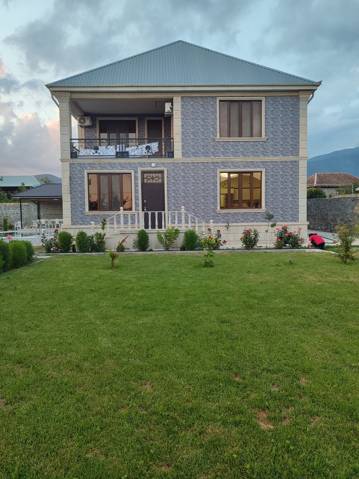 A two-story house features a patterned blue exterior and a light-colored roof. The front porch is adorned with decorative railings, while large windows allow natural light to enter. A neatly maintained lawn with shrubs and flowers surrounds the house, enhancing the outdoor appeal.
