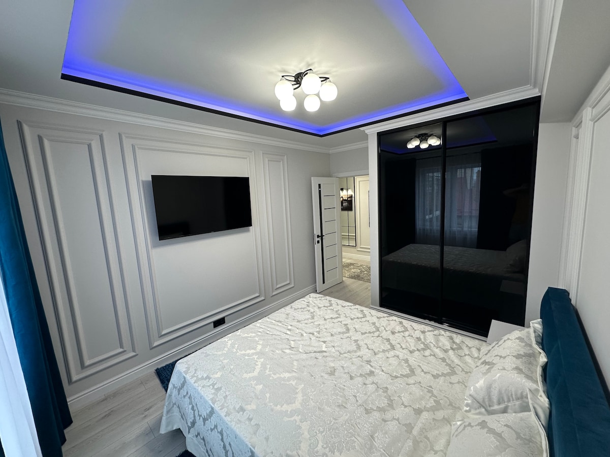 A modern bedroom features a spacious bed with a light-colored quilt. A sleek black wardrobe and a wall-mounted TV are positioned against intricately designed white walls. Soft blue lighting accentuates the ceiling, creating a calming atmosphere in the room.