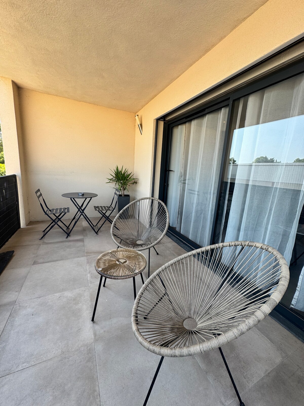 A spacious terrace is equipped with two round chairs and a small table, set against a backdrop of floor-to-ceiling glass doors. Natural light brightens the space, complemented by a potted plant, creating an ideal spot for relaxation and dining.