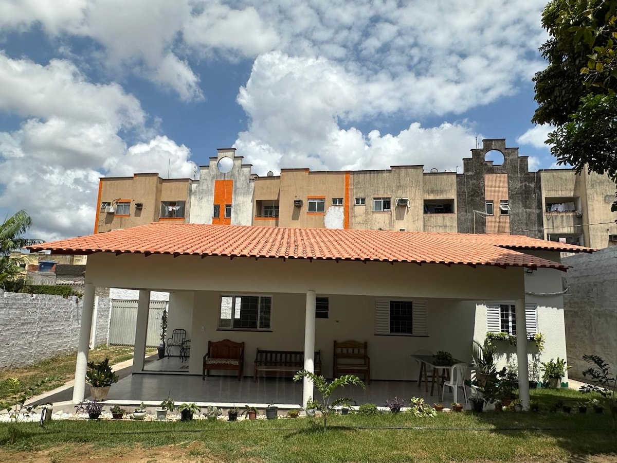 A covered outdoor area is presented, featuring a spacious porch with wooden benches and a tiled floor. Lush greenery and small plants are situated in the surrounding garden. In the background, modern apartment buildings rise against a partly cloudy sky.