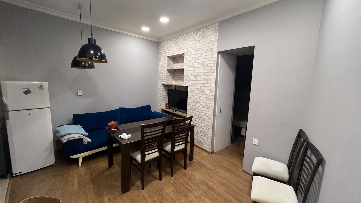 A modern living area features a blue sofa and a rectangular dining table surrounded by six wooden chairs. A flat-screen television is mounted on a white brick wall, enhancing the room's contemporary aesthetic. A door leads to a separate space, while a refrigerator is visible in the background.
