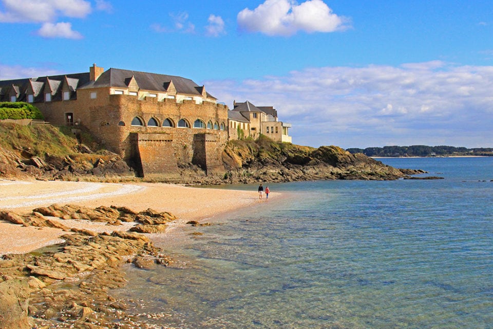 A coastline features a sandy beach bordered by smooth rocks and clear blue water, with two people walking along the shore. In the background, several buildings with sloped roofs are positioned along the edge of the beach, under a bright sky dotted with clouds.