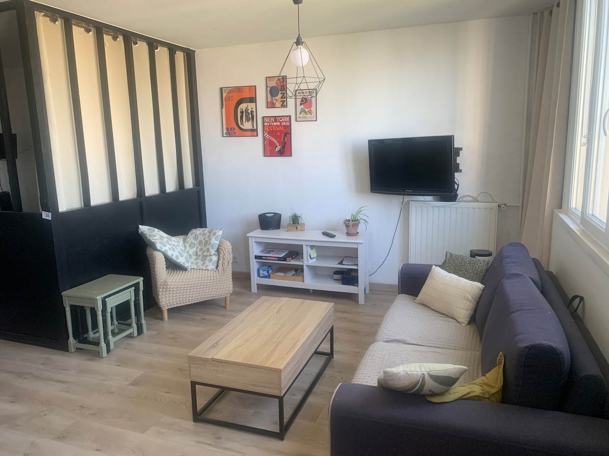 A comfortable living area features a dark sofa and a cozy armchair, arranged around a wooden coffee table. A small plant sits on the shelf below a wall-mounted television. Posters adorn the wall, while large windows allow natural light to fill the space.