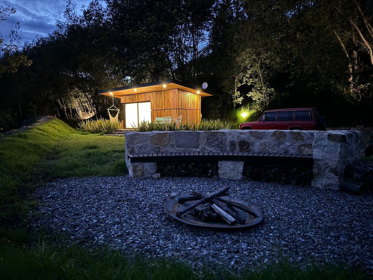 A cozy outdoor space is highlighted by a stone fire pit surrounded by gravel. A wooden tiny house is visible in the background, softly illuminated against the evening sky. Nearby, foliage and landscaping create a natural setting, enhancing the connection with the surrounding environment.