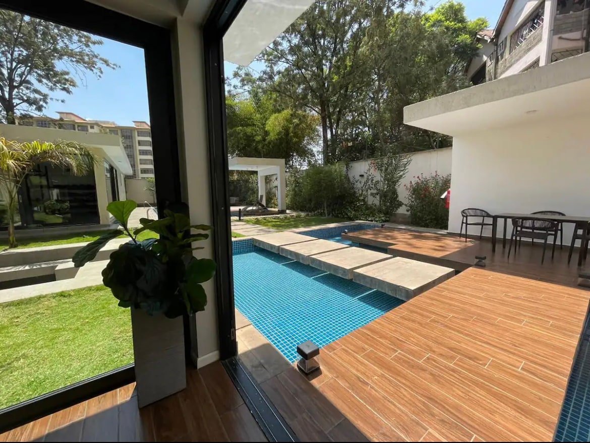 An indoor space opens to an outdoor area featuring a sleek pool. Sunlight reflects on the water, surrounded by manicured lawns and greenery. A dining area with chairs is visible adjacent to the pool, enhancing connection to the natural surroundings.