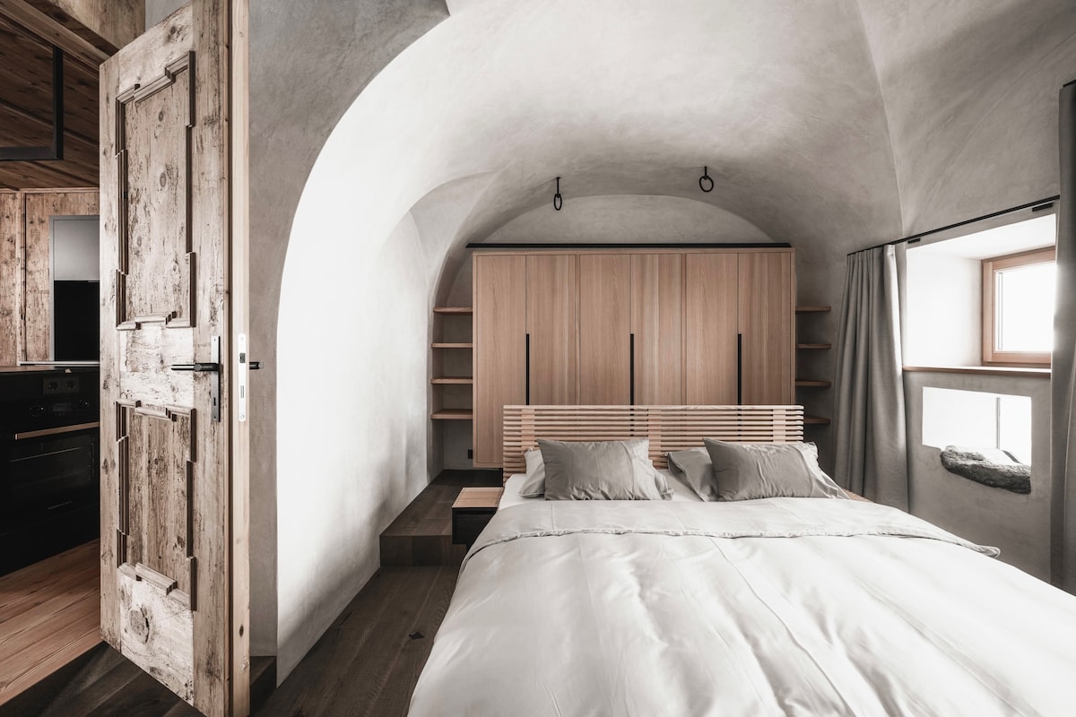 The bedroom features a spacious bed with soft bedding set against an arched ceiling. A wooden wardrobe is situated behind the bed, complemented by minimal shelves on either side. Natural light enters through a small window, enhancing the calm and serene environment.