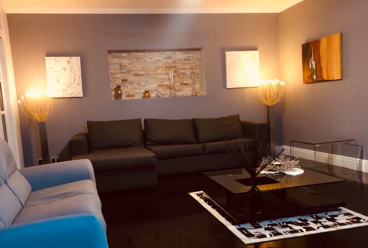 A modern living area is presented, featuring a charcoal gray sectional sofa and a matching armchair. Two abstract artworks adorn the walls, and an elegant glass coffee table sits at the center, complemented by a decorative arrangement. Soft lighting is provided by contemporary floor lamps.