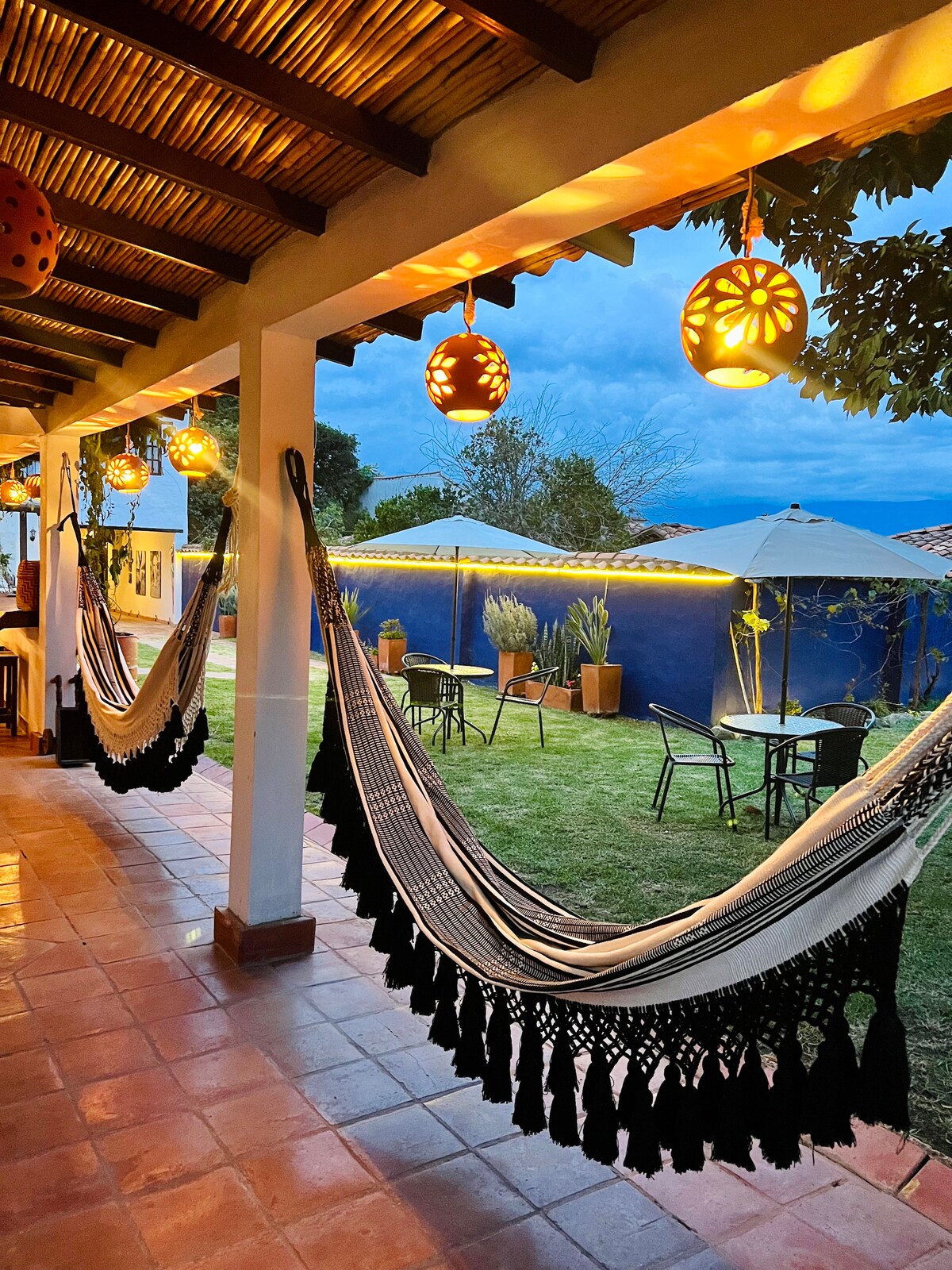An inviting outdoor space features several hammocks and small tables under the soft illumination of decorative lanterns. Lush greenery, including potted plants, surrounds the area, while two large umbrellas provide shade. A glimpse of the evening sky adds to the tranquil ambiance.