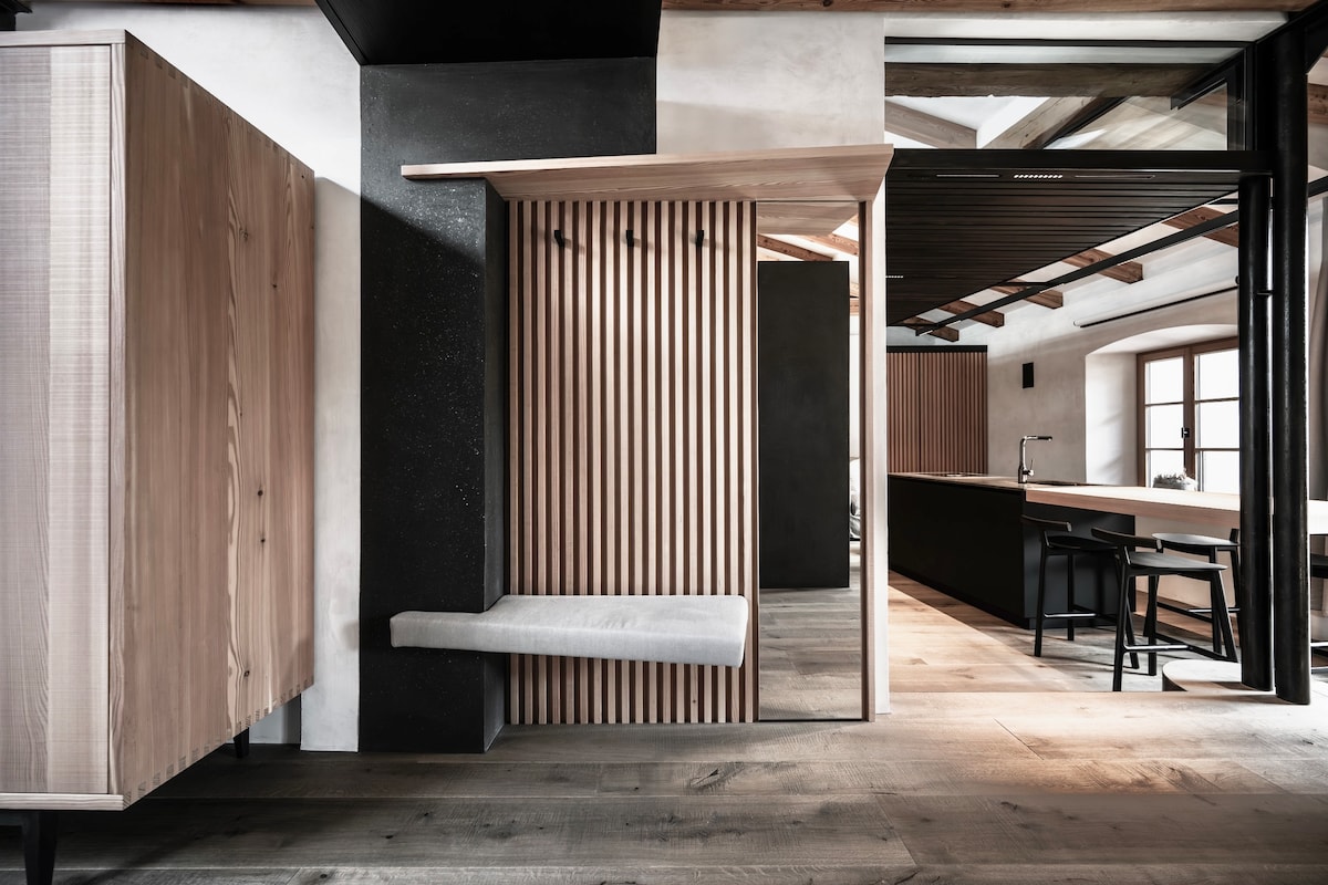 A modern entryway features a wooden bench and hooks set against a textured backdrop. Natural light filters through large windows, highlighting the clean lines of cabinetry and the spaciousness of the area. Sleek design elements combine functionality with a minimalist aesthetic.