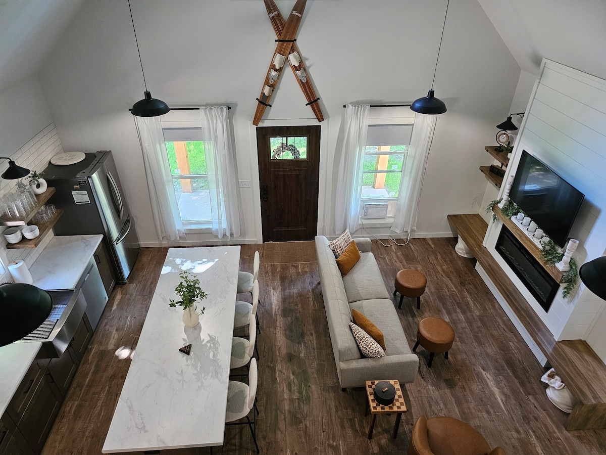 An open-concept living space features a spacious island table with seating for six. Natural light floods in from large windows, illuminating a light-colored sofa positioned near a fireplace. Decorative elements adorn the shelves, while pendant lights add a modern touch to the area.