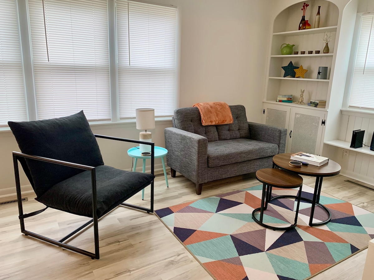 The living area features a cozy couch and an armchair positioned around a layered coffee table. A colorful geometric rug complements the space, while natural light enters through multiple windows adorned with light blinds. A lamp and decorative items are displayed on nearby shelves.