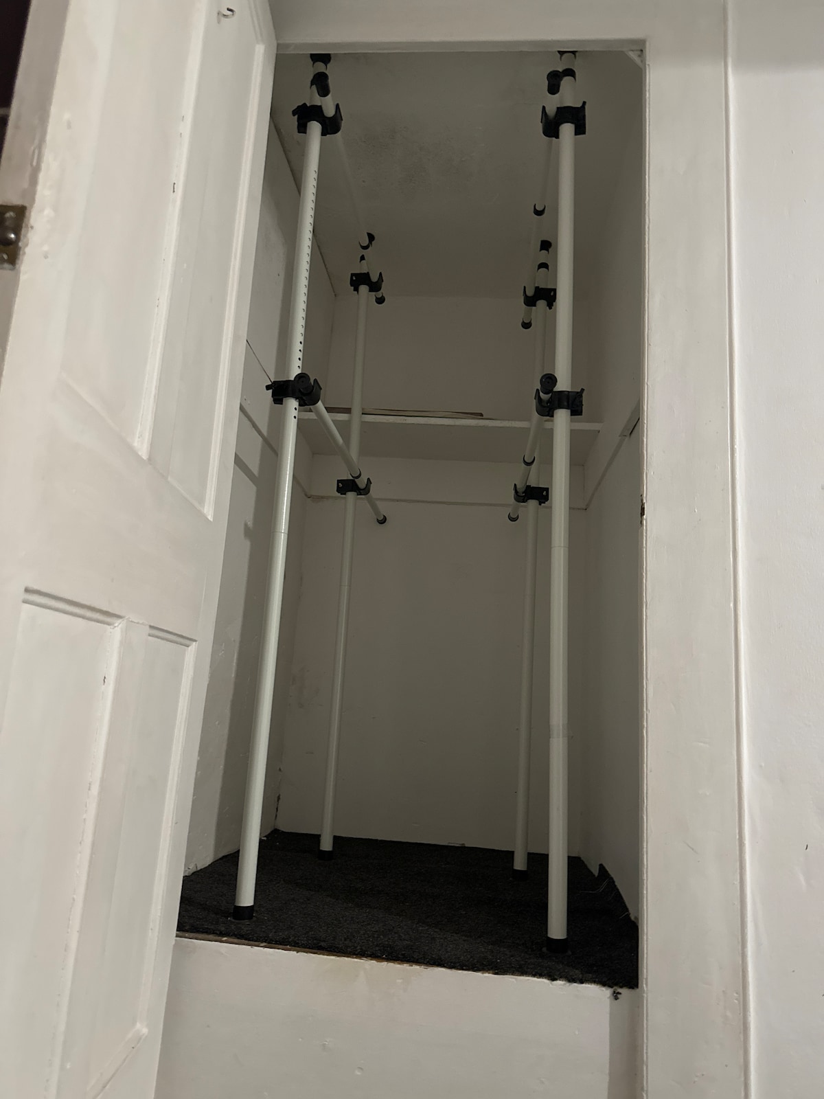 A simple closet is shown with white poles secured by black connectors, providing a space for hanging items. The floor is covered in dark carpet, and a white door remains open, revealing the closet's interior.