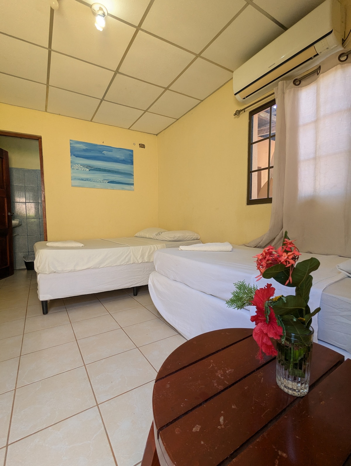 A modest guest room features three spacious beds with white linens arranged thoughtfully. A small table holds a decorative vase with tropical flowers. Natural light enters through a nearby window draped with a light curtain, illuminating the room's soft yellow walls.