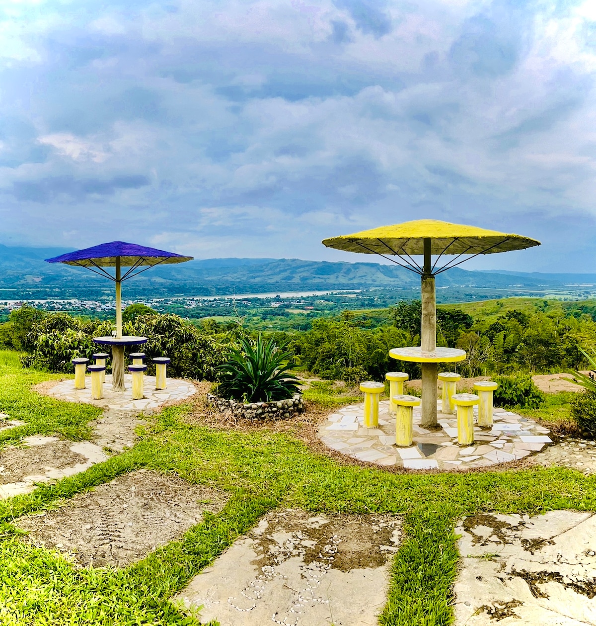 Two outdoor seating areas are positioned on a grassy hilltop, each shaded by large umbrellas in contrasting colors of blue and yellow. The tables and stools are circular and multi-colored, surrounded by unobstructed views of the valley and distant mountains.