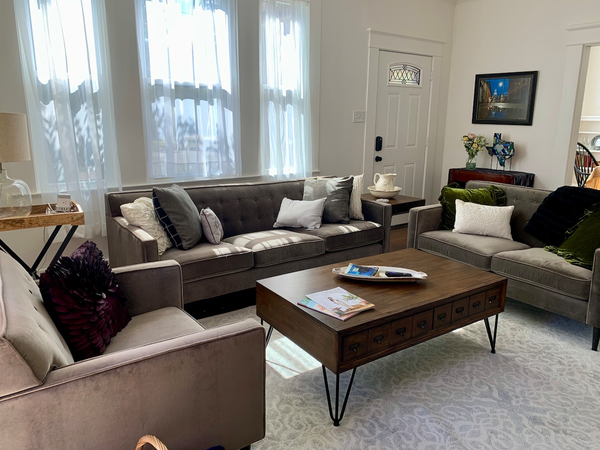 A bright living room features large windows adorned with sheer curtains, allowing natural light to fill the space. Two comfortable sofas face each other across a coffee table, which holds magazines. Decorative pillows enhance the sofas' inviting appearance, creating a relaxed atmosphere.
