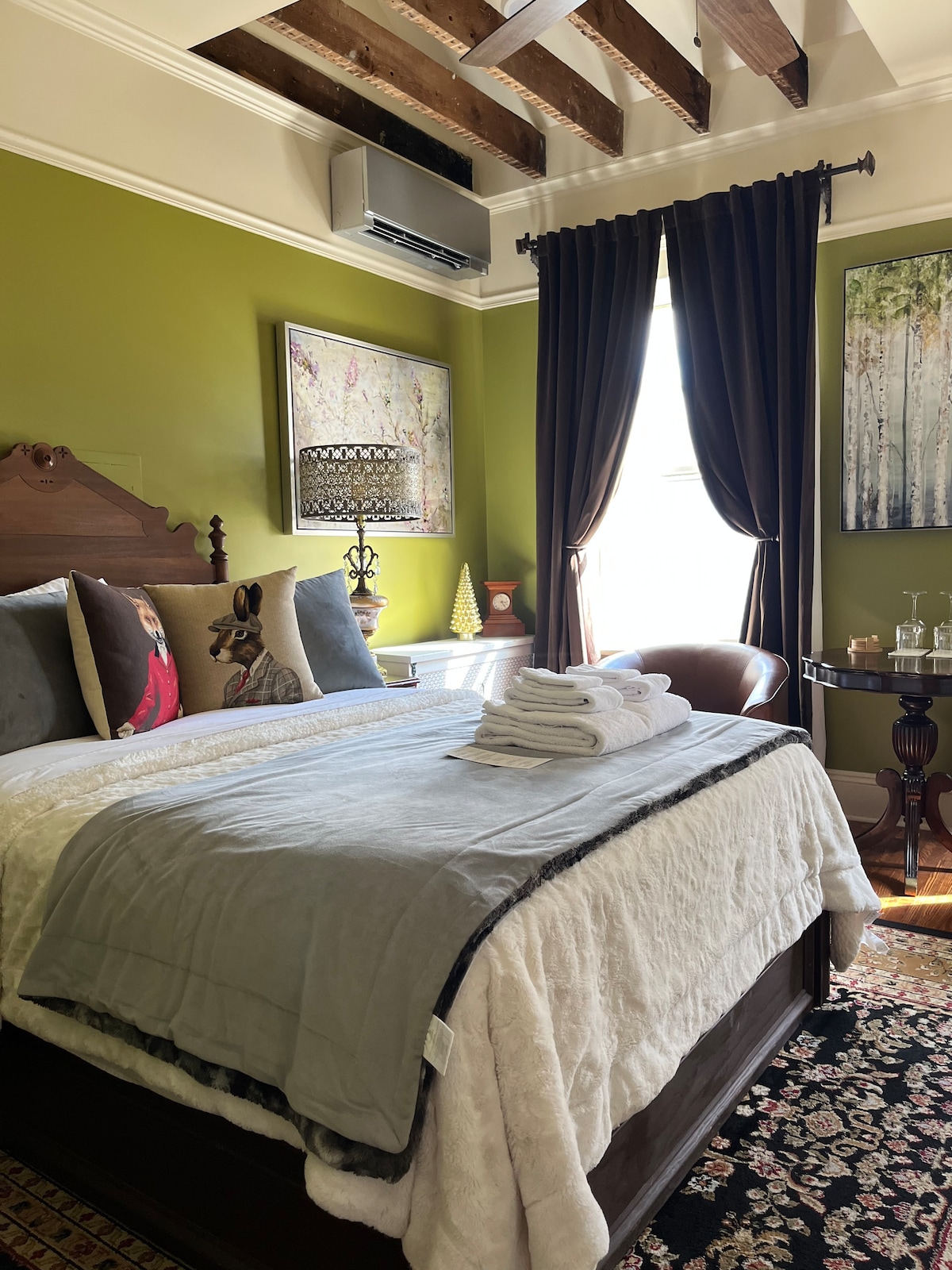 The room features a queen size bed dressed in soft linens, with neatly folded towels displayed on the bed. Rich green walls are complemented by dark curtains and wooden beams overhead. A cozy chair and side table are positioned near a window, allowing natural light to enter.
