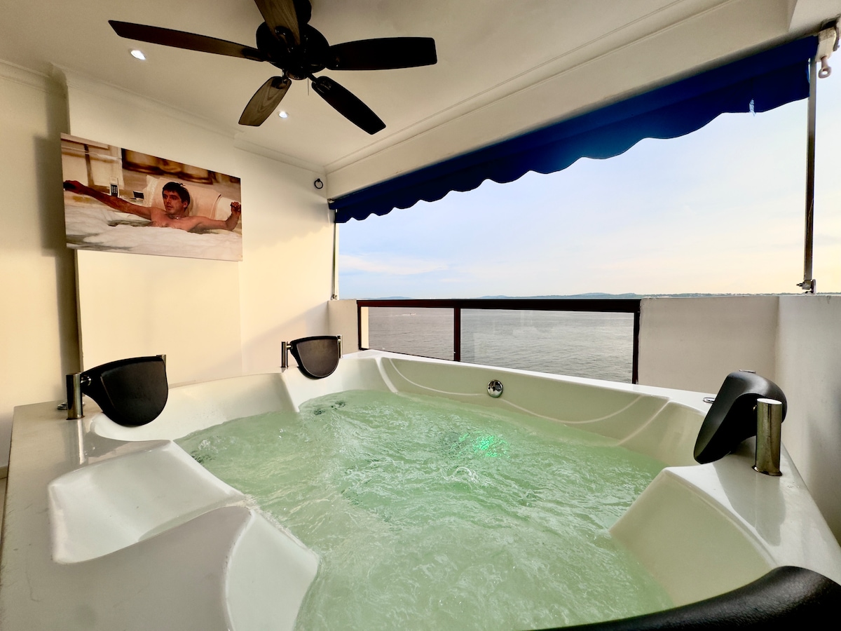 A private jacuzzi designed for four is situated on a balcony, overlooking ocean views. The jacuzzi features two built-in seats with backrests. Above, a ceiling fan is mounted, providing airflow. A flat-screen television is mounted on the wall, and a blue awning offers shade.