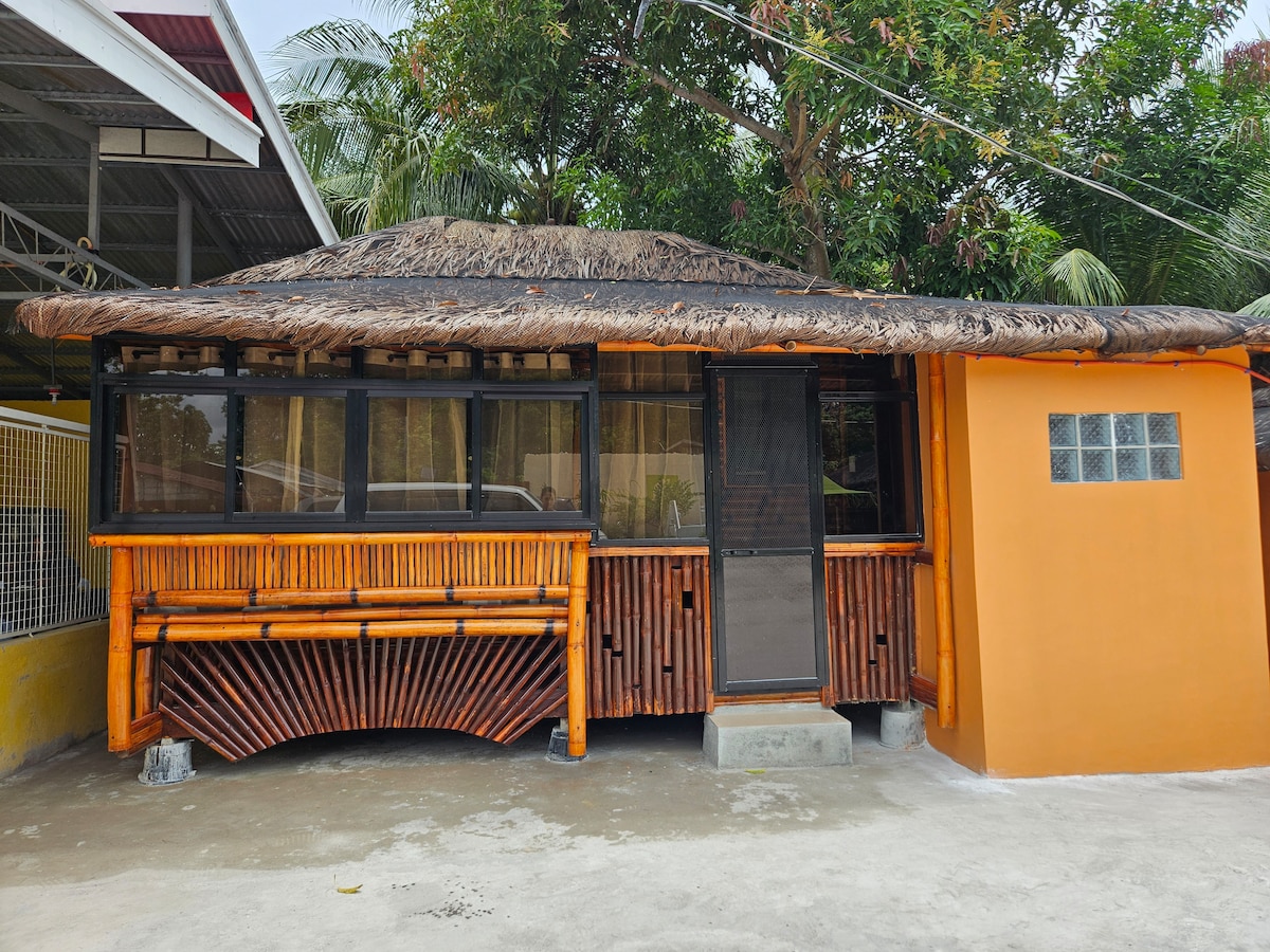 A colorful exterior features a bamboo structure with a thatched roof. Large windows invite natural light, while the vibrant orange wall complements the surrounding greenery. A concrete pathway leads to the entrance, emphasizing the welcoming nature of the home.