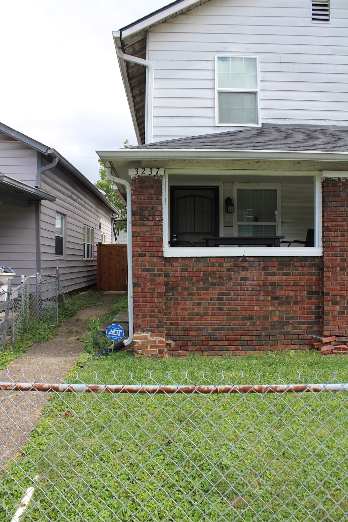 A white duplex is shown with red brick accents around the porch area. A small path leads up to the entrance, while a chain-link fence outlines the front yard, which features green grass. A nearby wooden fence is visible on the left side.