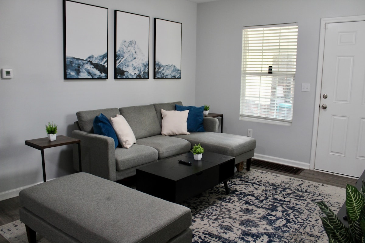 A modern living room is displayed, featuring a light gray sectional sofa adorned with blue accent pillows. A sleek black coffee table is placed in front, surrounded by a decorative rug. Three framed mountain prints enhance the walls, while a window allows natural light to enter.