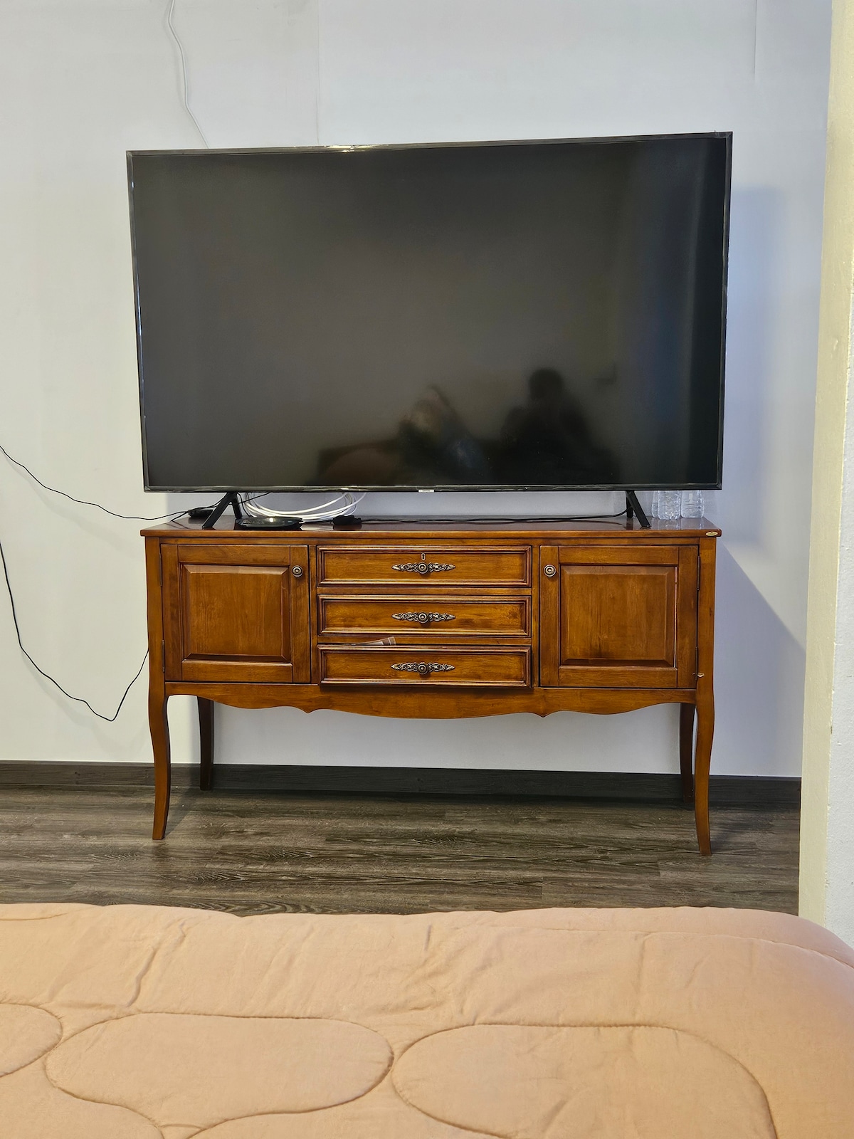 A large flat-screen TV is mounted on a wall above a wooden console with multiple drawers. The console features a classic design with curved legs, positioned against a plain backdrop, while the floor displays a dark wood finish.