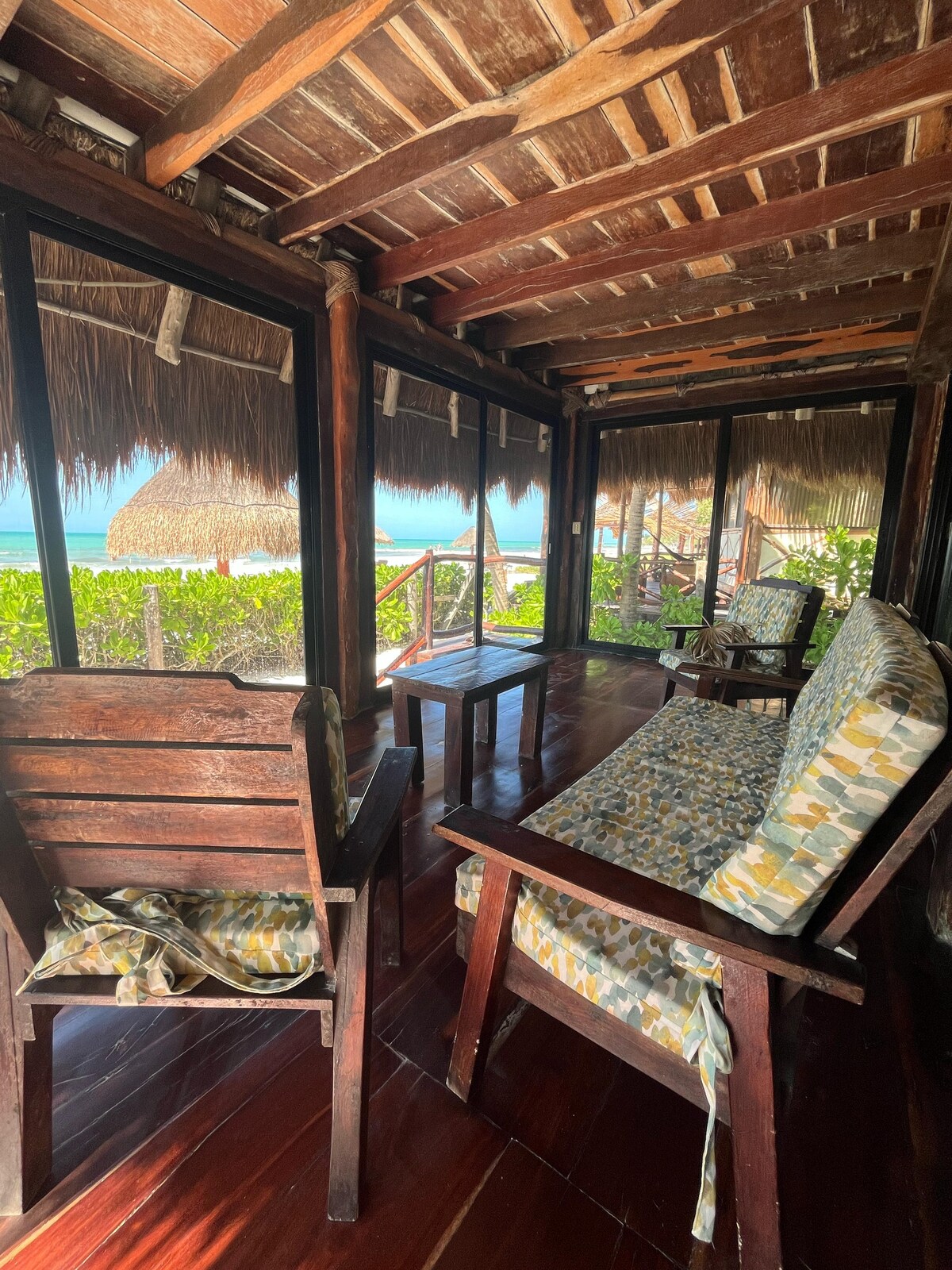A cozy seating area is depicted with two wooden chairs and a small table, adorned with cushions featuring a vibrant pattern. The space is enclosed by glass walls, providing views of the lush greenery and beach beyond. Natural light fills the area, enhancing the warm wood tones.