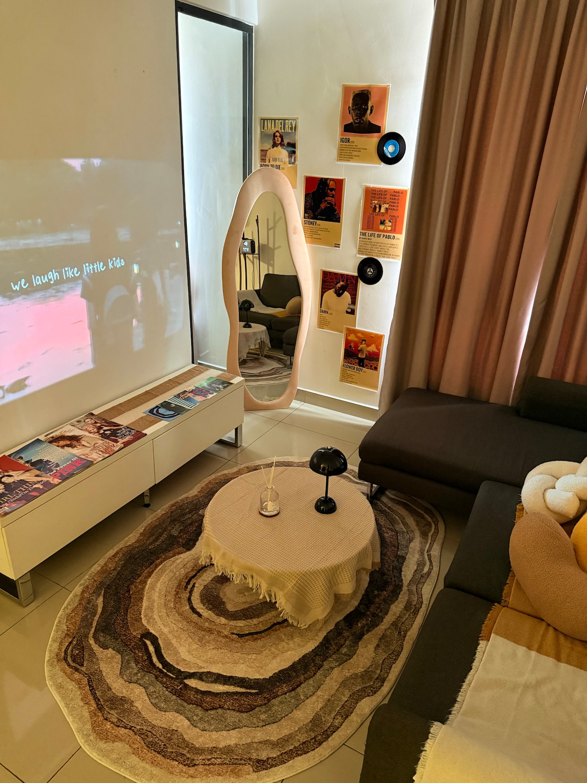 A cozy living area features a neutral-toned sofa and a round coffee table placed on a textured rug. A large mirror reflects the space, and a projector screen displays a movie. Decorative posters and vinyl records adorn the walls, enhancing the relaxed vibe.