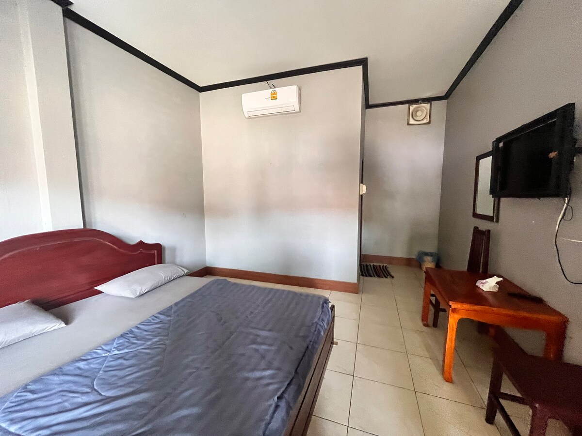 A simple sleeping area is presented with a spacious bed dressed in neutral linens. A small wooden table and a chair are positioned against the wall. A mounted television and an air conditioning unit are visible, enhancing comfort in the room.
