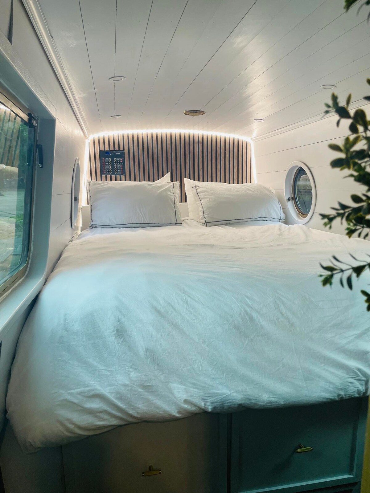 A spacious sleeping area features a king-size bed adorned with crisp white bedding. Two large cushions rest against a headboard with wooden slats, while soft lighting creates a calming ambiance. Circular windows allow natural light to illuminate the space.