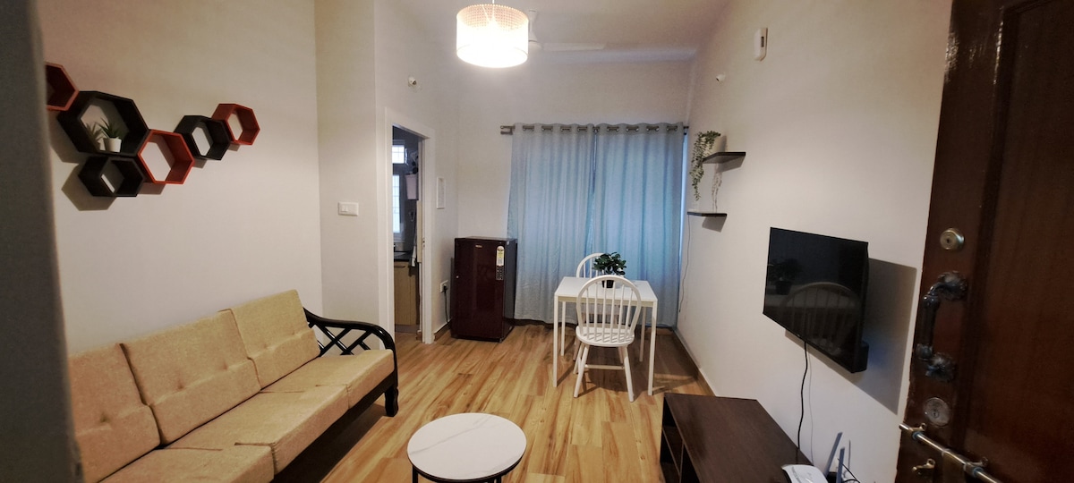 A welcoming living area features a beige sofa against a light-colored wall. A small round coffee table sits in front, and a white dining table is positioned beside a window with sheer curtains. Wall decor includes hexagonal shelves with plants, and a television is mounted nearby.