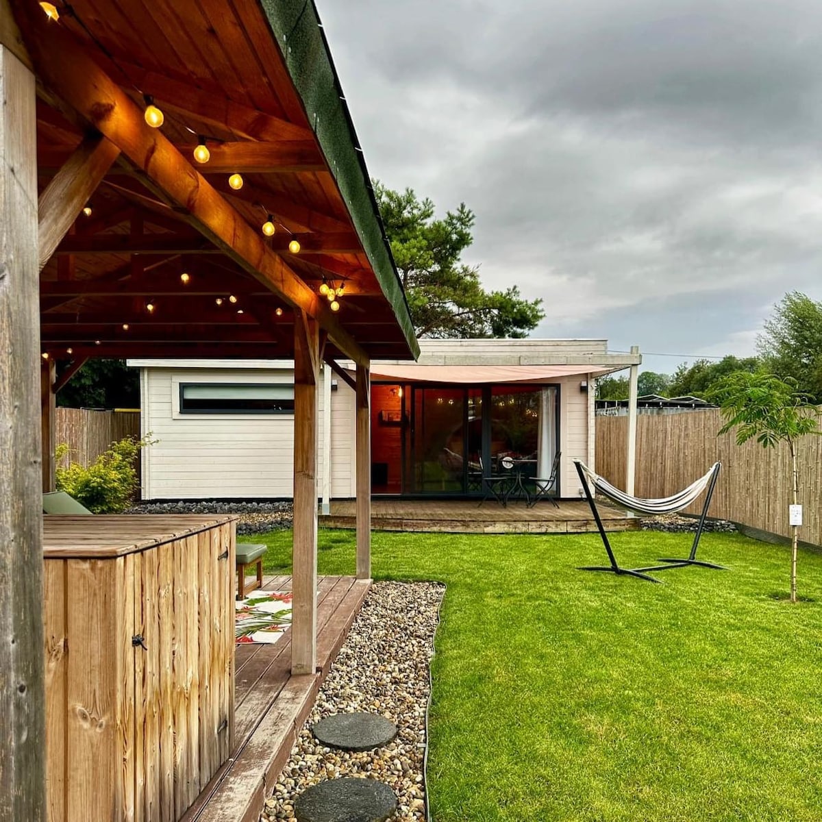 A charming outdoor area is featured, showcasing a wooden pavilion with string lights. A grassy lawn surrounds a relaxing hammock and a small decorative tree, while a modern cottage is visible in the background, framed by a wooden fence.