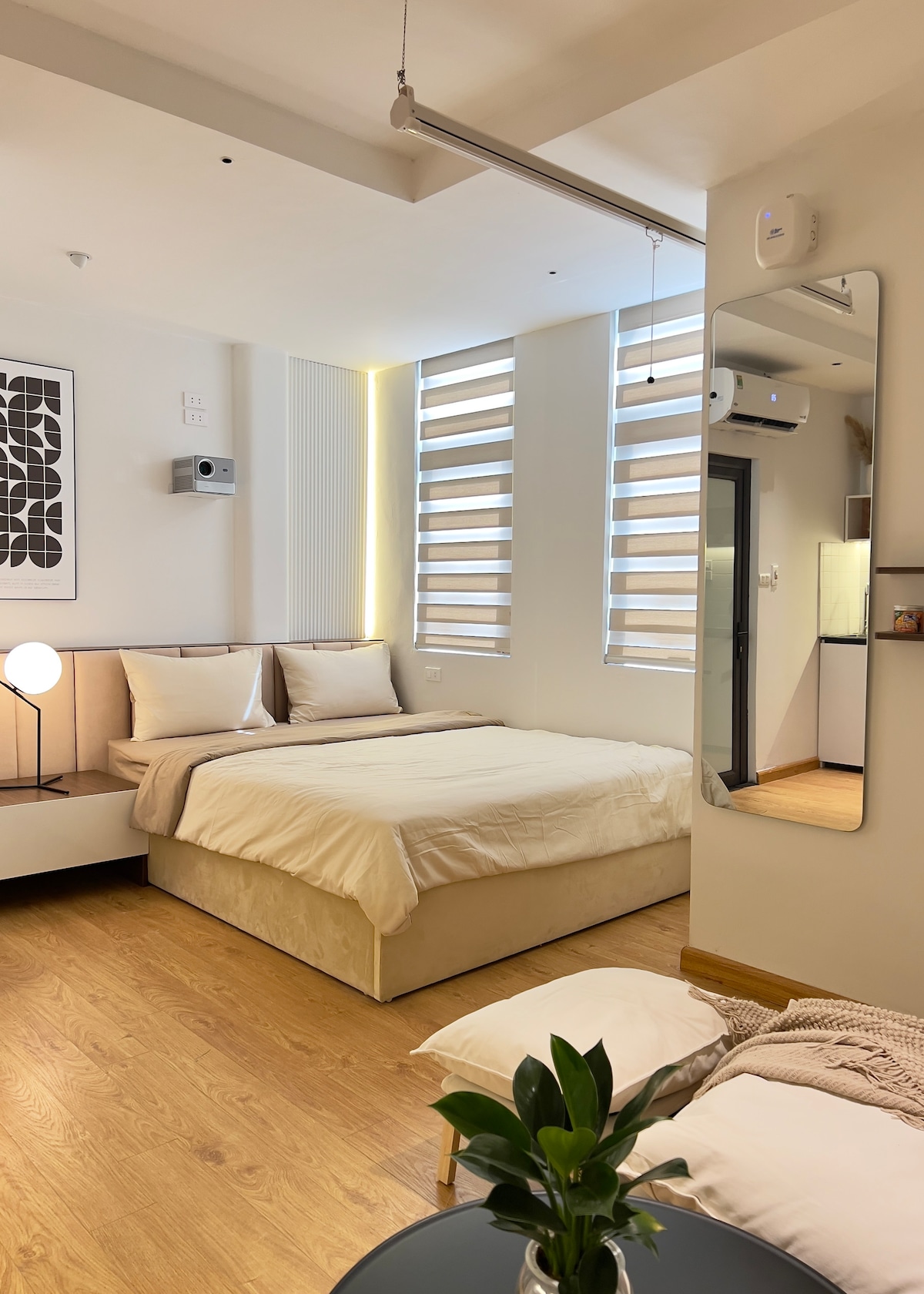 The studio room features a spacious bed adorned with neutral-colored linens. Two small windows allow natural light to fill the space, complemented by a minimalist mirror and a bedside lamp. A kitchenette is visible in the background, emphasizing the room's functionality.
