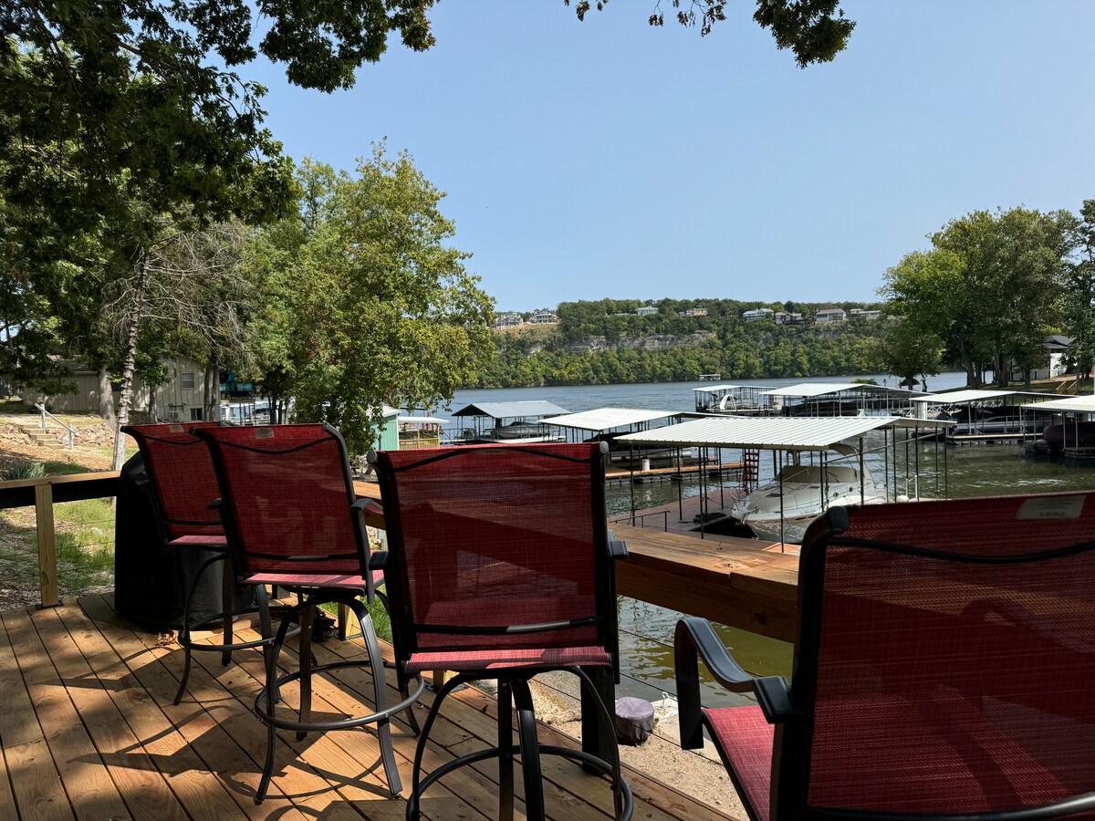 A spacious outdoor deck is presented, featuring several red chairs positioned for lake views. The calm water is bordered by lush green trees, while nearby boat docks are visible against a backdrop of rolling hills under a clear sky.