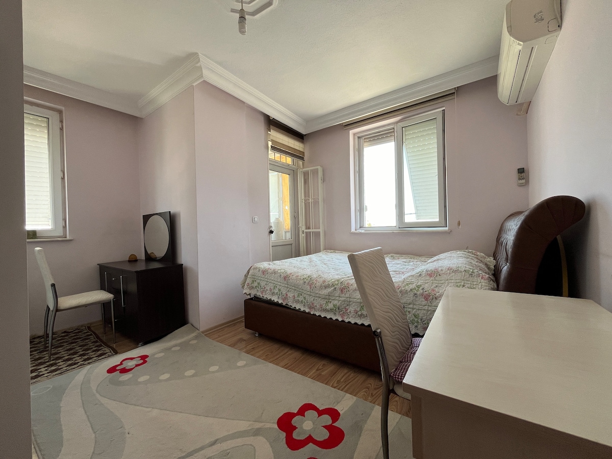 A softly painted bedroom features a spacious bed with a floral covering. A modest desk, mirror, and chair are positioned in the room. Bright light enters through two windows, which are framed by subtle window treatments, contributing to the calming atmosphere.