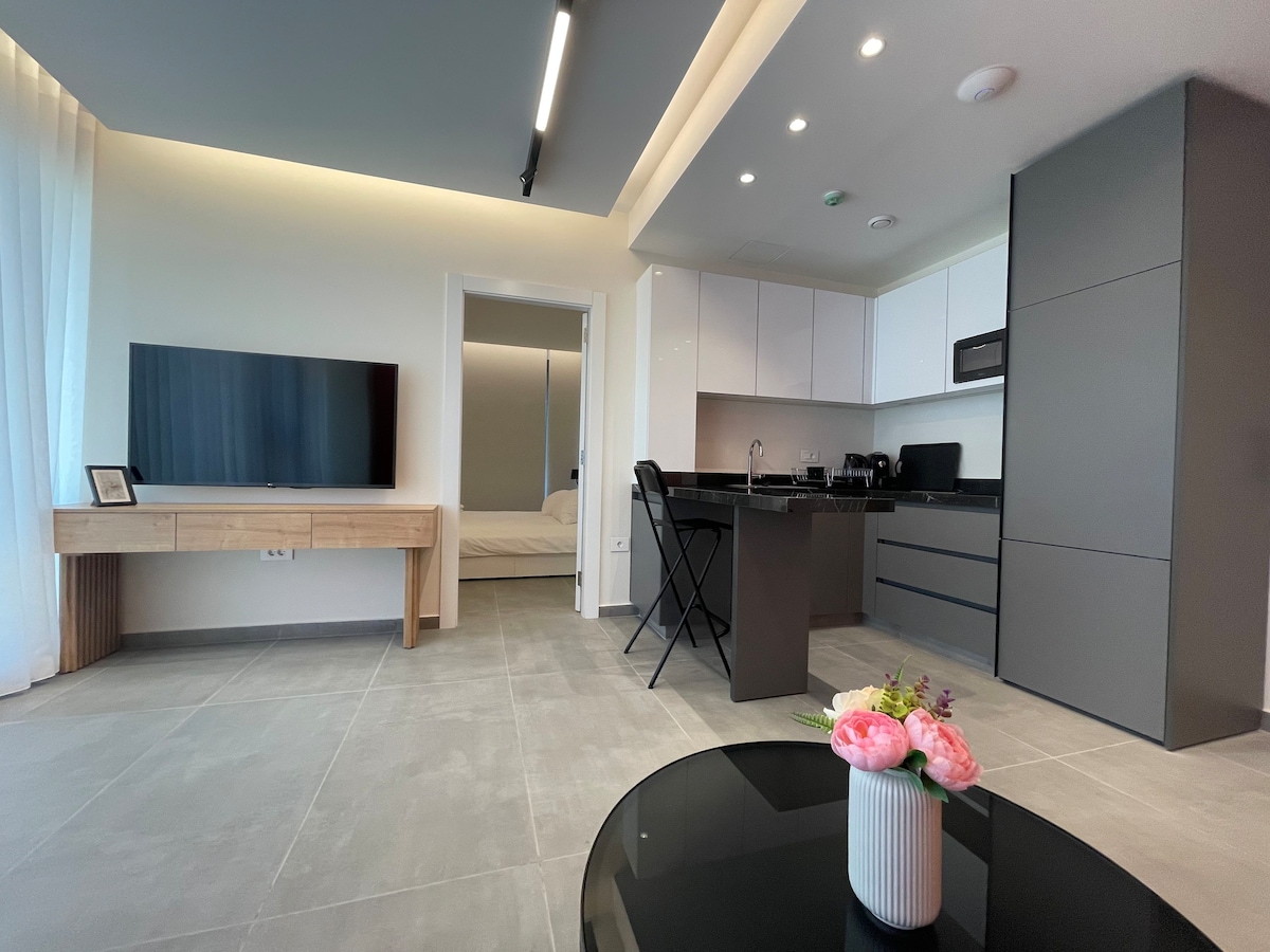 The open-plan living area features a modern kitchen with sleek black cabinetry and essential appliances, including a stove and fridge. A cozy dining space with two black chairs complements a flat-screen TV mounted on the wall. Natural light fills the room through large windows.