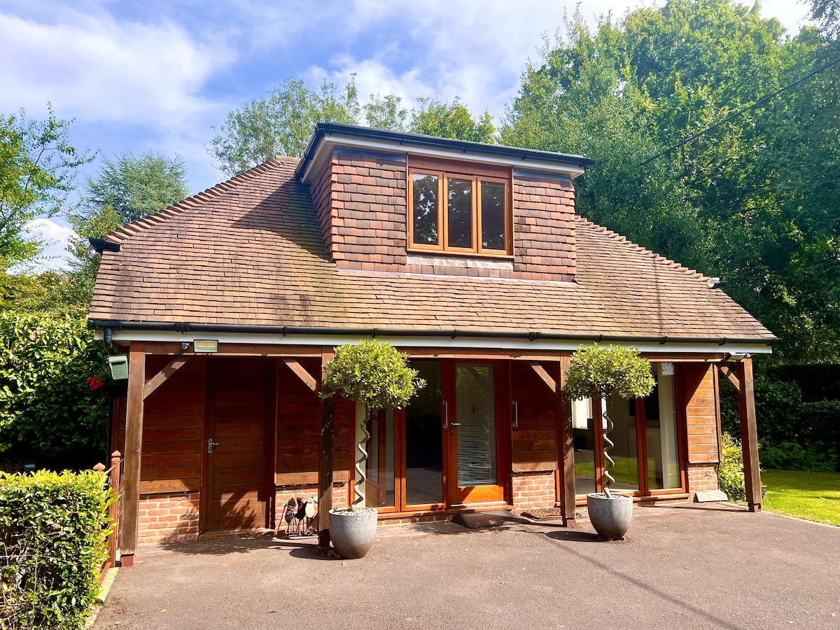 The exterior of the detached Annexe is shown, featuring a wooden facade with a sloped roof and large windows. A covered entrance is supported by columns, while neatly trimmed hedges and two decorative planters with shrubs add greenery to the setting.