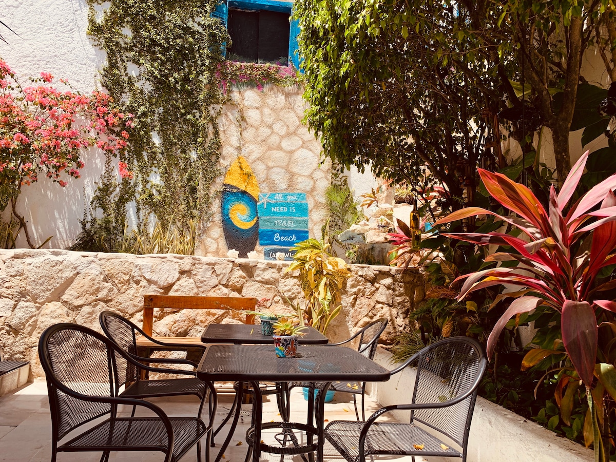 An outdoor patio area is featured, furnished with several black metal chairs arranged around dark tables. Colorful plants and a stone wall create a serene backdrop. A blue sign decorated with a wave motif adds a touch of charm to the space, surrounded by lush greenery.