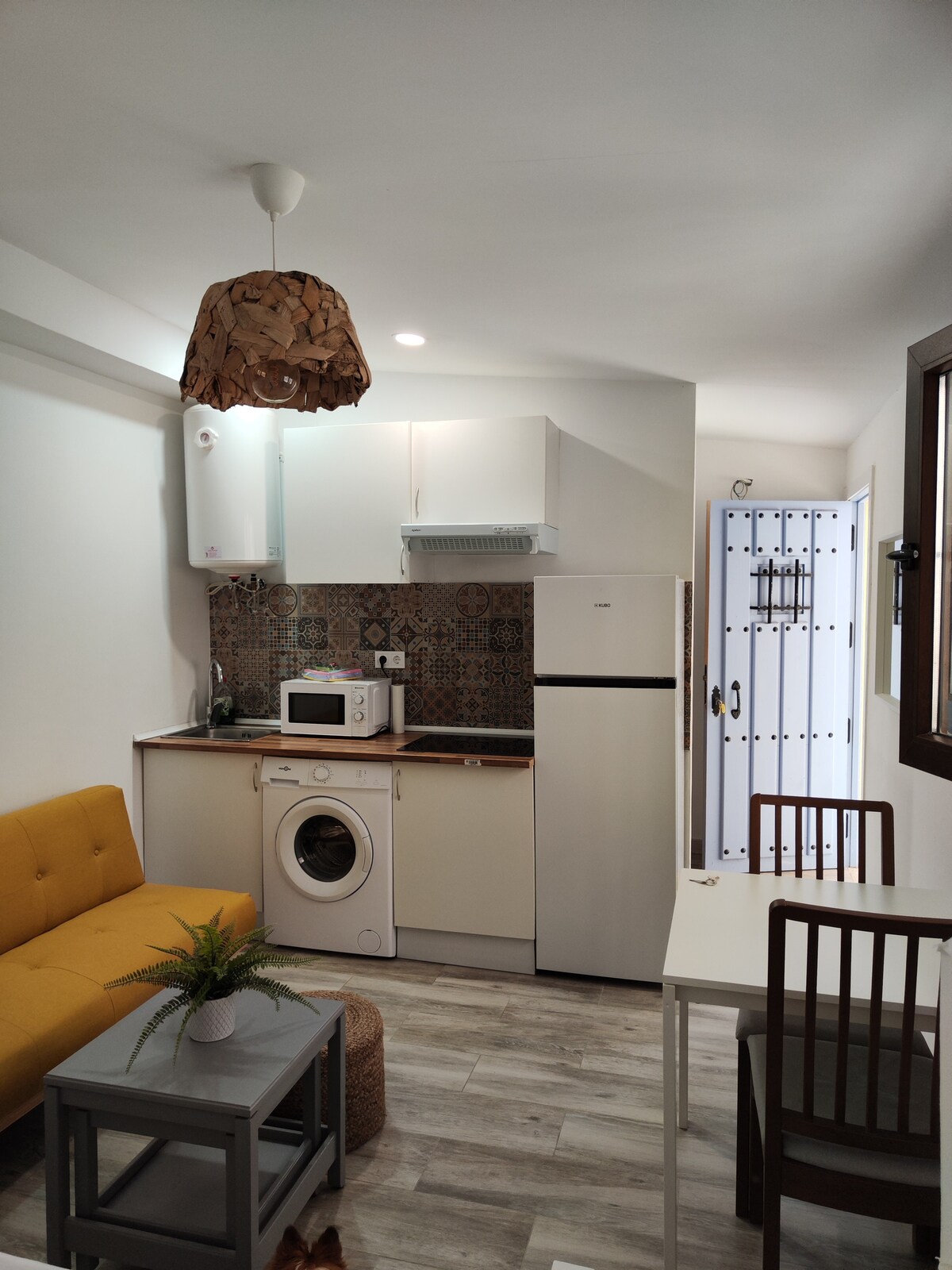 A compact kitchenette is equipped with a refrigerator, microwave, and a washing machine. A cozy yellow sofa complements the space, while a small dining table with two chairs is positioned nearby. Natural light enters through the window, enhancing the inviting atmosphere.