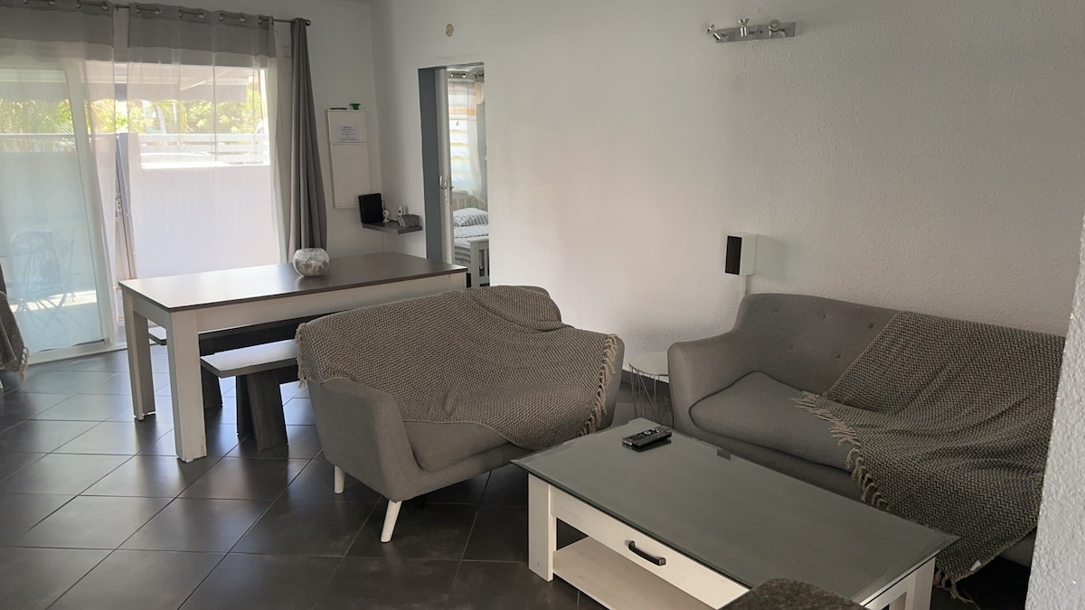 A light-filled living area features a pair of gray sofas, one draped with a soft blanket. A sleek coffee table sits in the center, with a remote placed on it. A simple dining table is positioned to the side, complemented by sliding glass doors that lead outside.