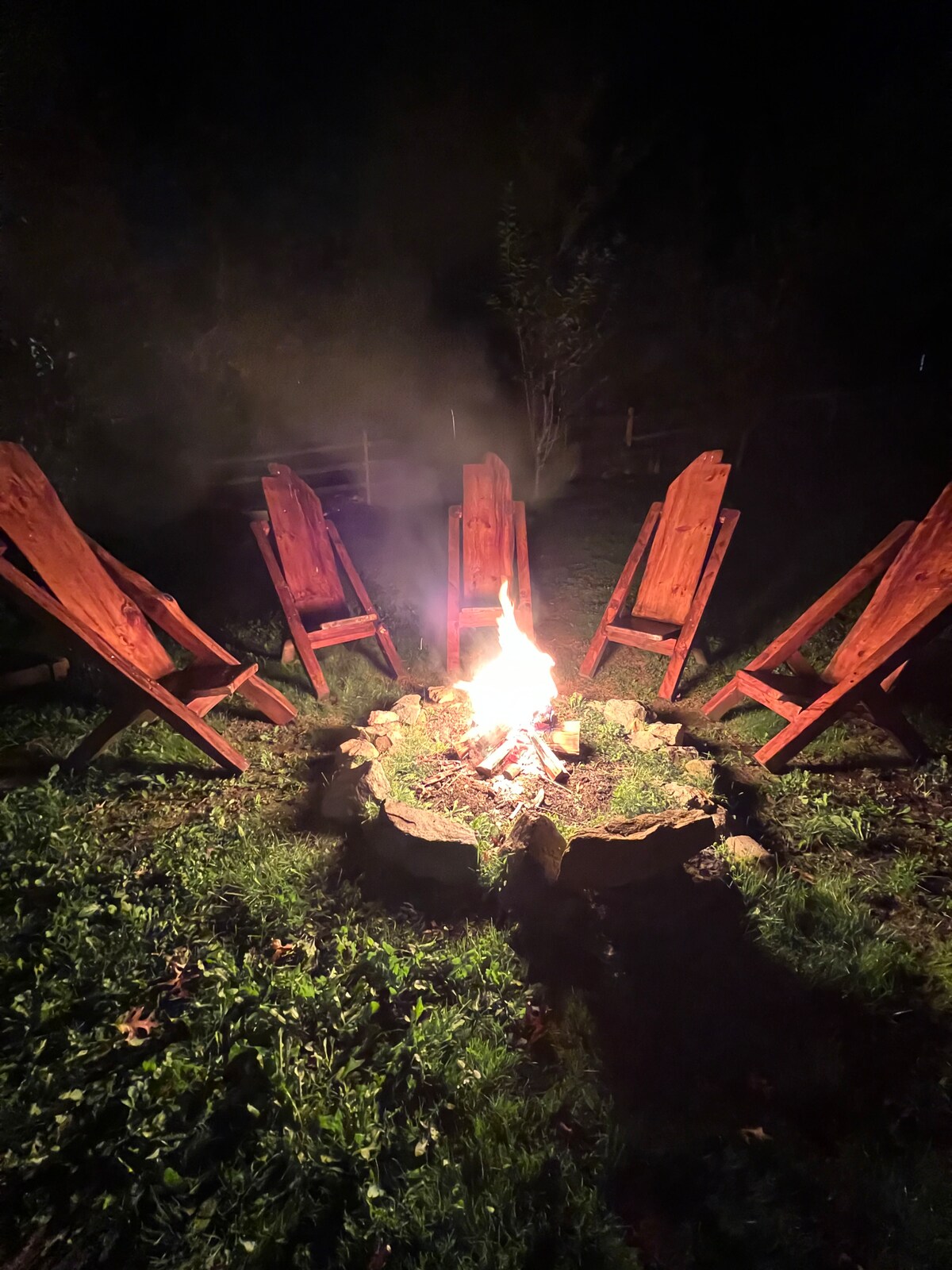 A fire pit surrounded by five wooden chairs is illuminated by the flickering flames. The grassy area surrounding the pit adds a natural setting, while trees provide a backdrop, creating a cozy gathering space for evenings outdoors.