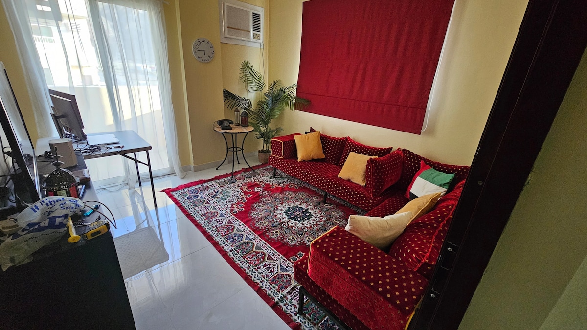 A cozy lounge area features a rich red sofa set adorned with decorative cushions. A traditional patterned rug covers the floor, and large windows allow natural light to illuminate the space. A small table and a plant add to the inviting atmosphere.